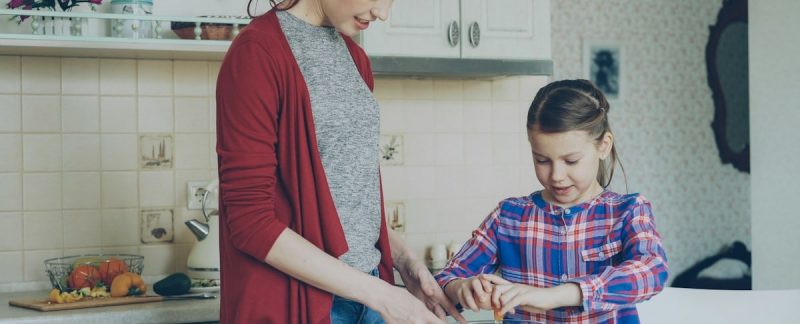 Little cheerful girl helping her mother in the kitchen cropping egg into bowl and mixing dough for cookies. Family, food, home and people concept